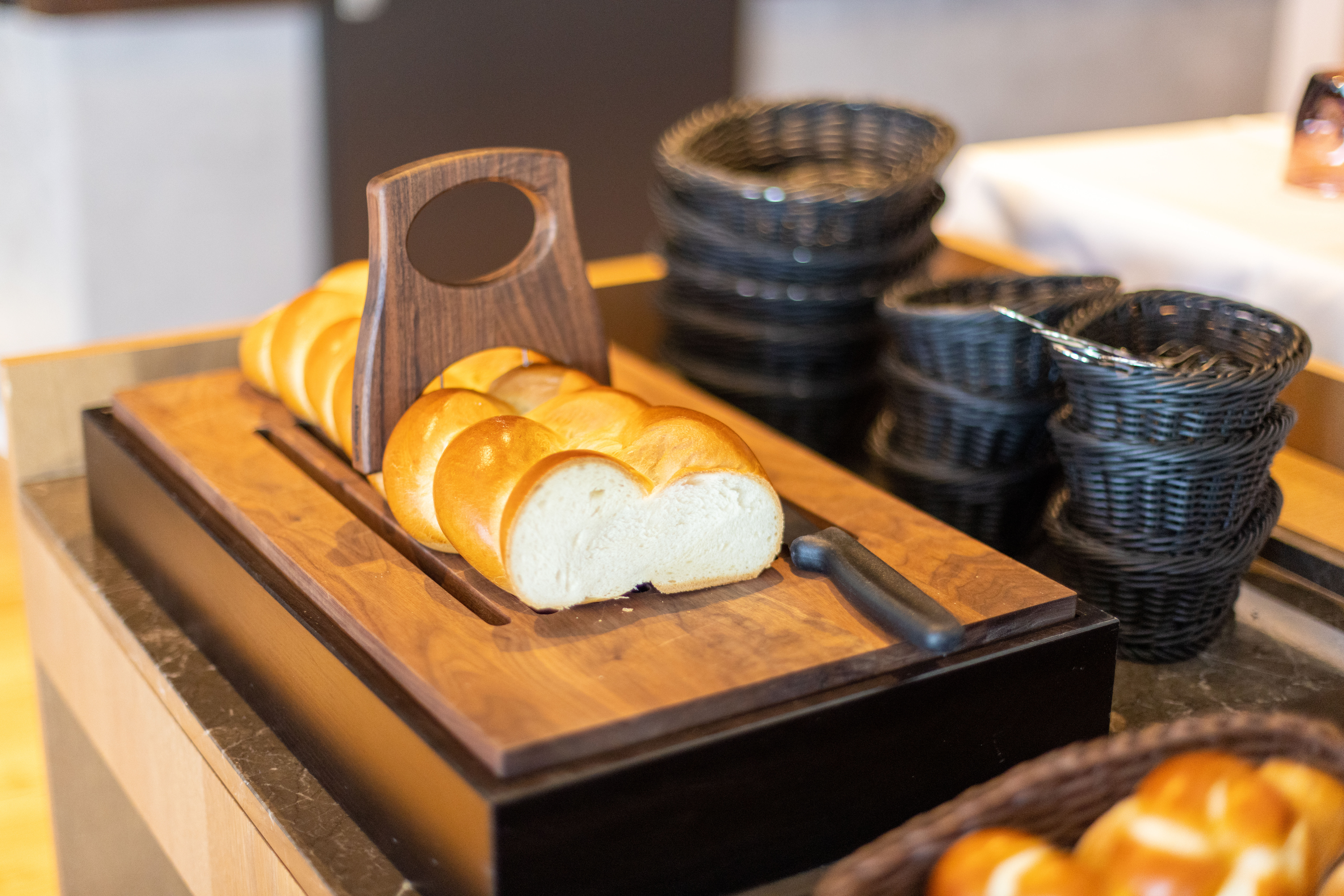 Nahaufnahme vom Zopf auf einem Holzbrett mit Griff und einem Messer daneben. Im Hintergrund sind gefüllte Brotkörbe zu sehen. Das Bild ist Teil vom Macardo Brunch Angebot.